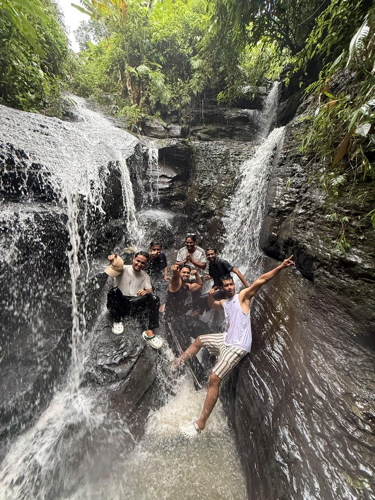 Aehtesham and friends cheering under a waterfall in a green, rocky gorge